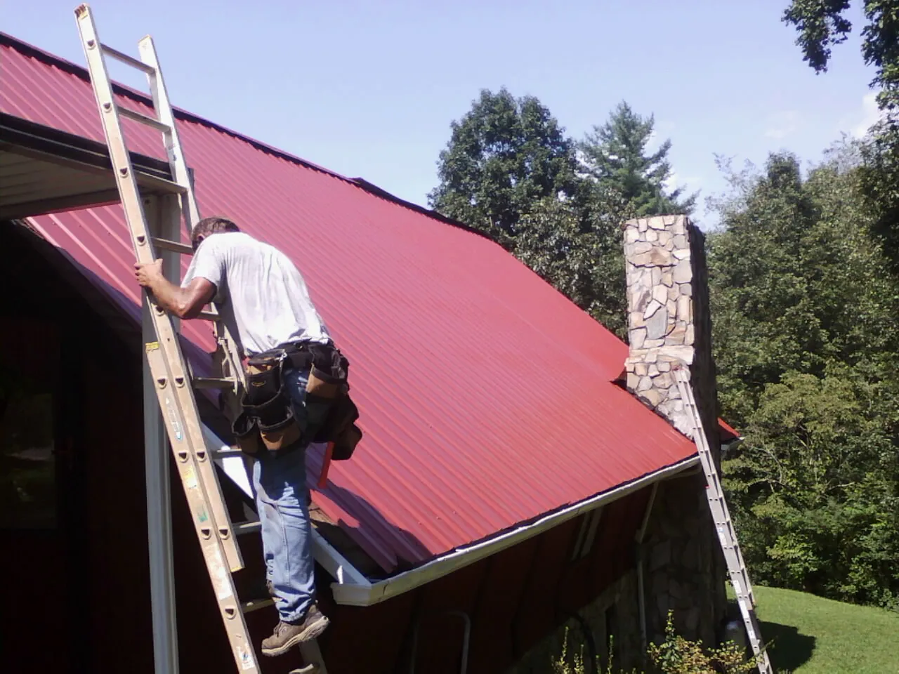 Roofer on a ladder beside a red metal roof and stone chimney — Soffit & Fascia Repair by Roofing Brothers in Harbert
